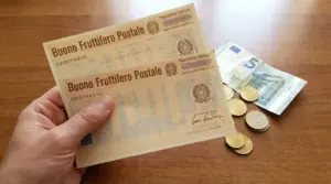 Mano che tiene buoni fruttiferi postali con monete e banconota da cinque euro su tavolo in legno