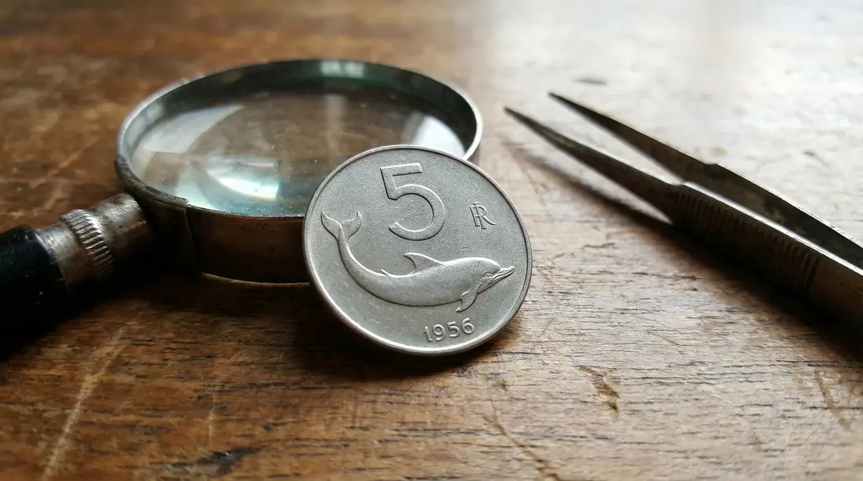 Vecchia moneta da 5 lire con delfino del 1956 accanto a lente d'ingrandimento e pinzette su tavolo di legno