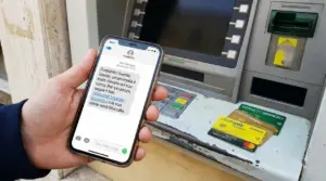 Una persona tiene uno smartphone con un messaggio truffaldino davanti a un bancomat con carte bancarie